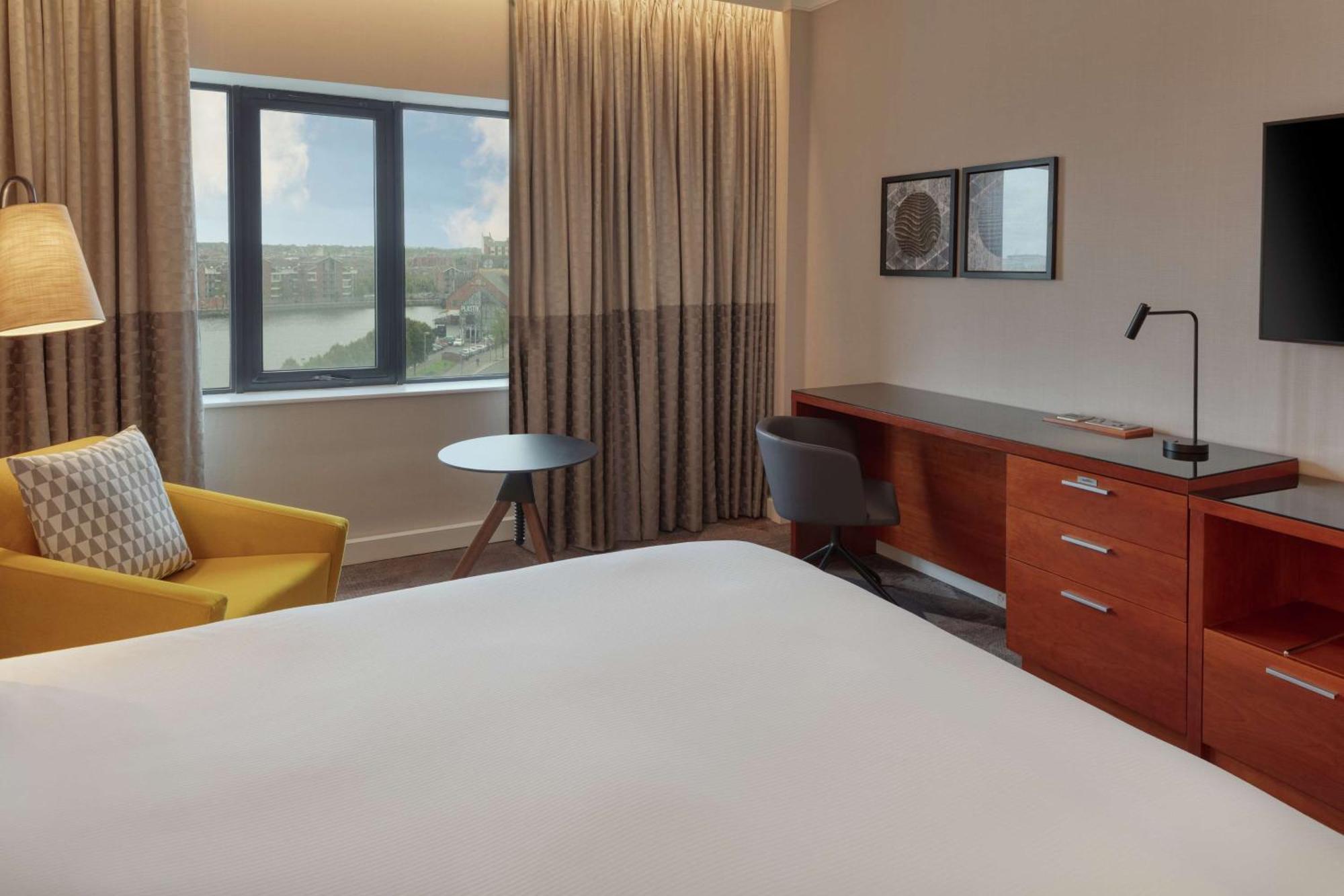 Hilton Belfast Hotel Екстериор снимка The photo shows a modern hotel room. It features a large bed with a plain white bedspread, and there's a stylish yellow armchair next to it. A small round table is placed nearby, along with a gray chair. The room has large windows covered with light 