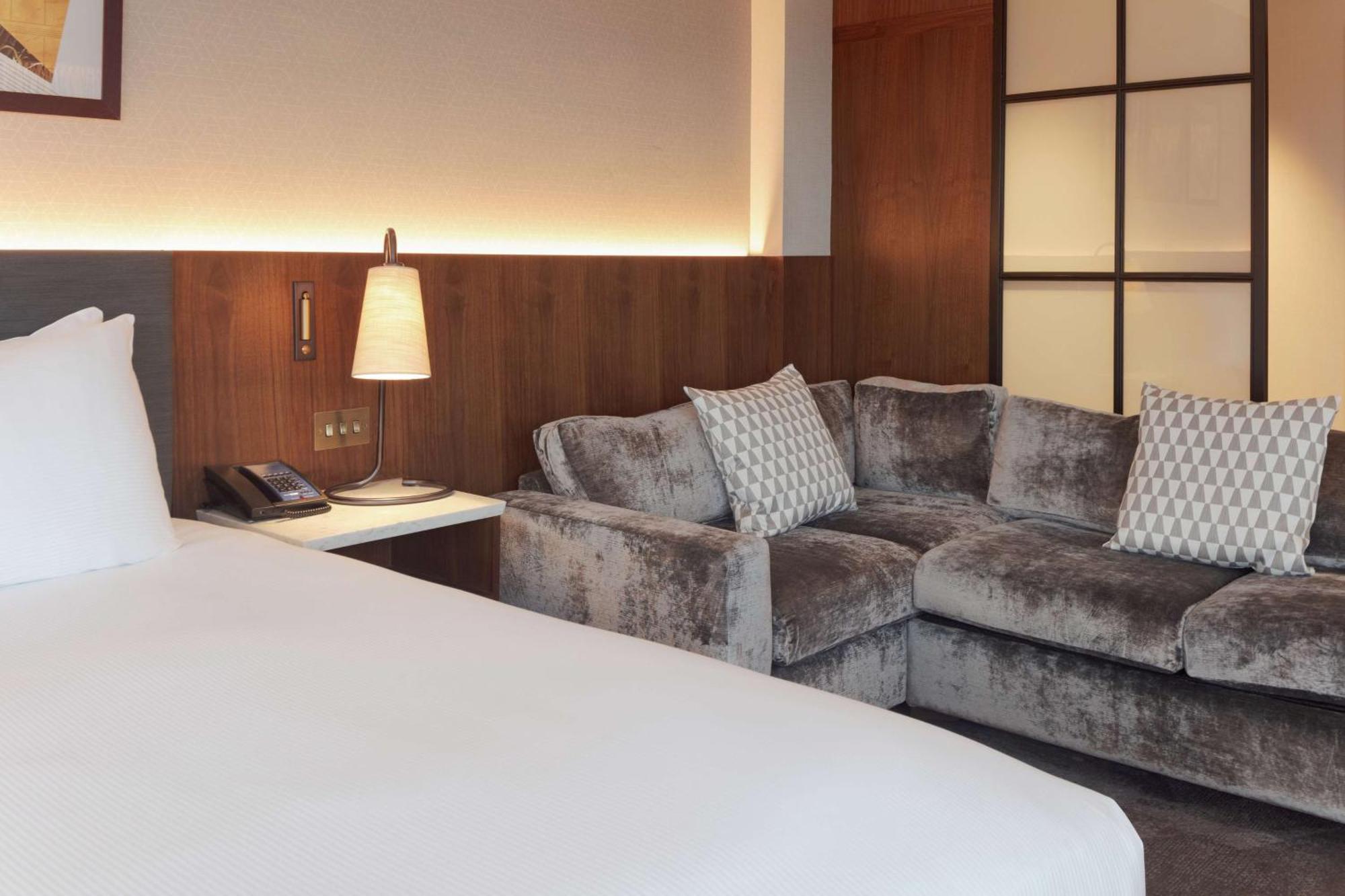 Hilton Belfast Hotel Екстериор снимка The photo shows an interior room, likely a hotel or a modern living space. In the foreground, there's a neatly made bed with white bedding. To the side, there is a plush sofa in shades of gray, adorned with decorative pillows of a similar color. Next