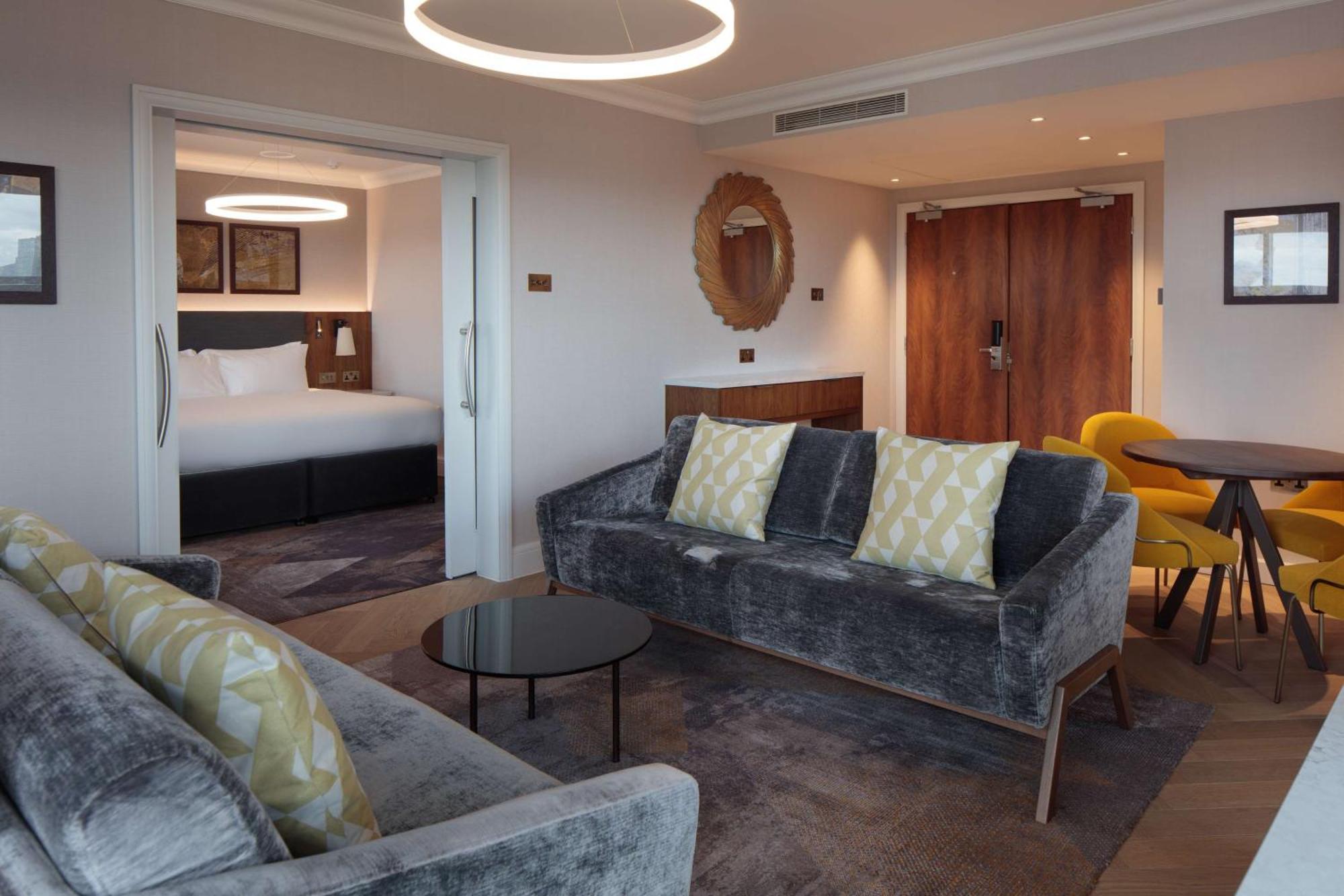 Hilton Belfast Hotel Екстериор снимка The photo shows a modern and stylish hotel suite. In the foreground, there is a comfortable gray sofa with decorative pillows and a small black coffee table. Adjacent to the sofa is a dining area with a round table and yellow chairs. In the backgroun