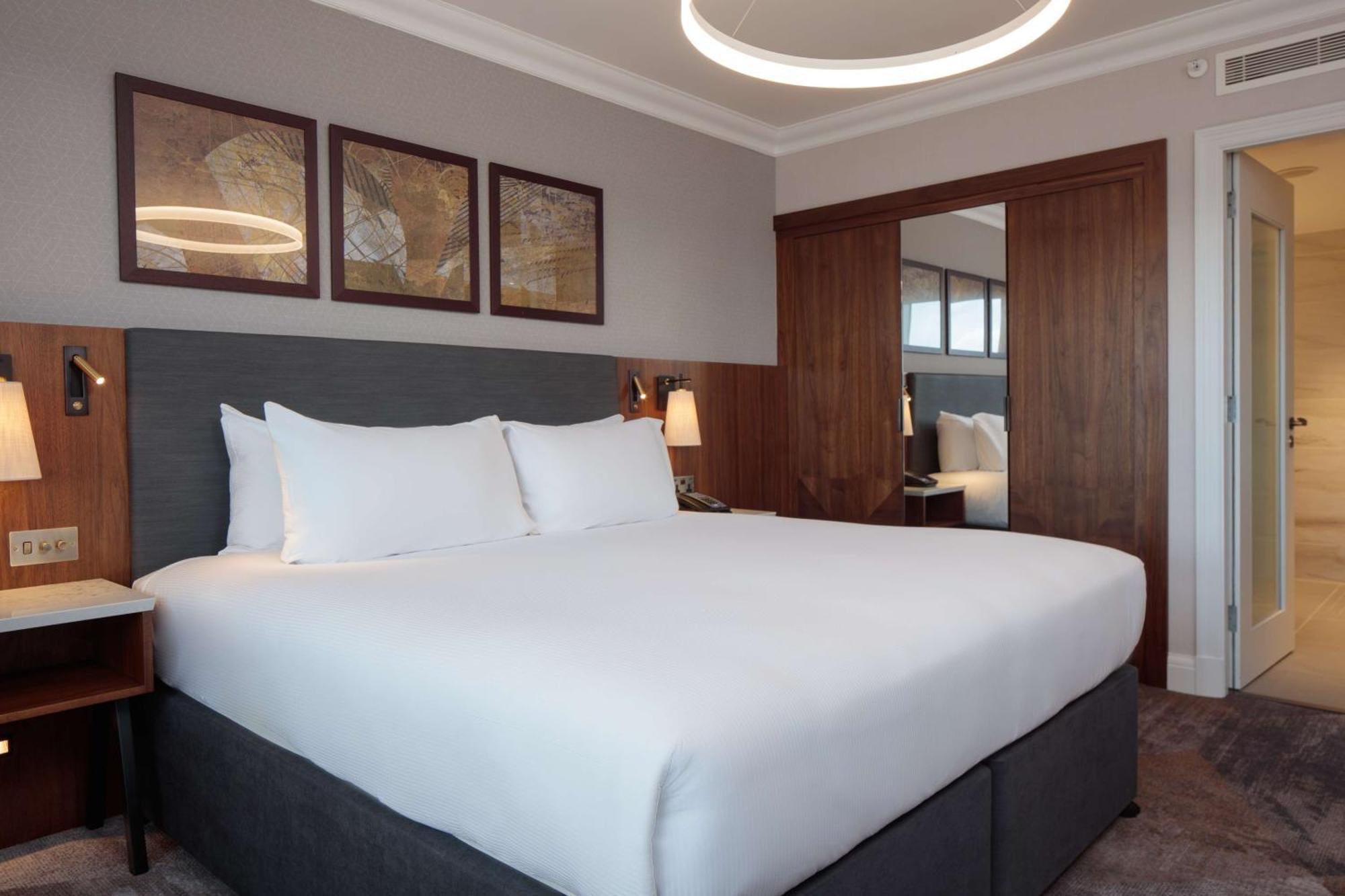 Hilton Belfast Hotel Екстериор снимка The photo shows a modern hotel room with a clean and contemporary design. The focal point is a large bed with crisp white bedding and two fluffy pillows. The headboard is gray, and next to the bed, there are stylish bedside lamps. Above the bed, ther