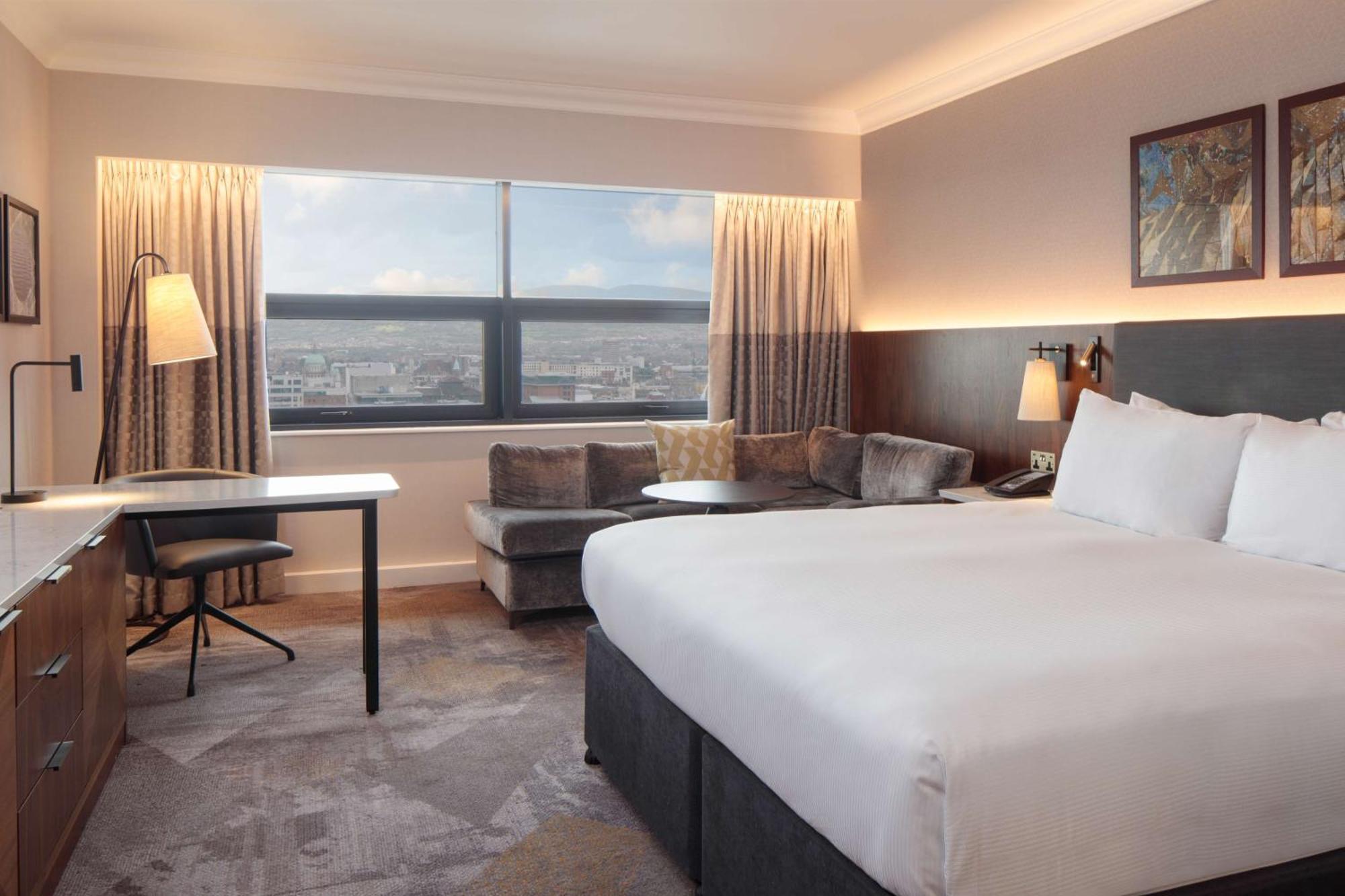 Hilton Belfast Hotel Екстериор снимка The photo shows a modern hotel room featuring a large bed with white linens positioned against a wall. On one side, there is a small seating area with a couch and a round table. A desk and chair are also present, suggesting a workspace. The room has 