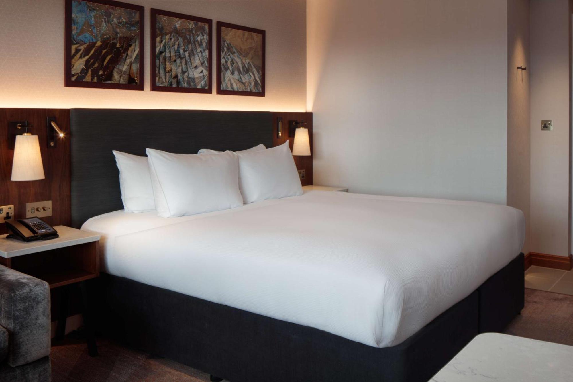 Hilton Belfast Hotel Екстериор снимка The photo shows a modern hotel room featuring a large bed with white bedding and several pillows. The bed is positioned against a dark-colored headboard. On the wall above the bed, there are three framed artworks. The room has soft lighting, and ther