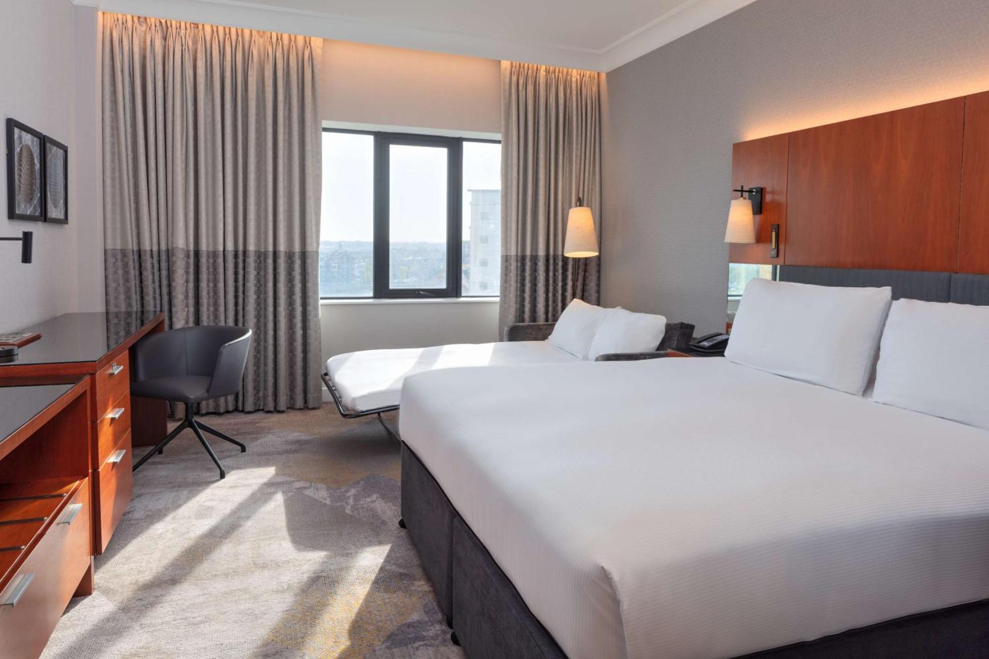 Hilton Belfast Hotel Екстериор снимка The photo shows a modern hotel room. It features two beds with white linens and a simple, elegant design. There is a large window allowing natural light to enter, covered by light-colored curtains. A comfortable chair and a small desk are placed near