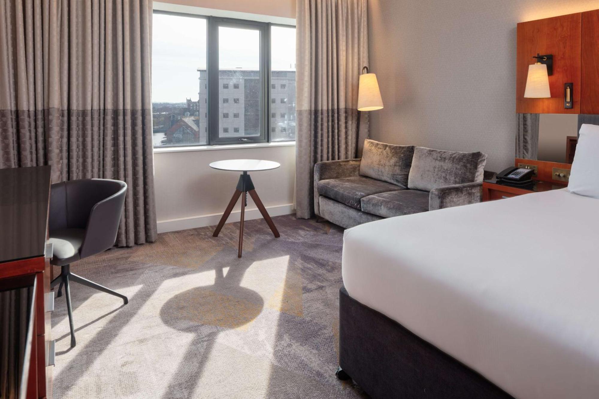 Hilton Belfast Hotel Екстериор снимка The photo shows a hotel room featuring modern furnishings. There is a large bed with white bedding positioned against one wall. Near the window, which has light-colored curtains, there is a small round table with a tripod-style base. A sleek gray sof