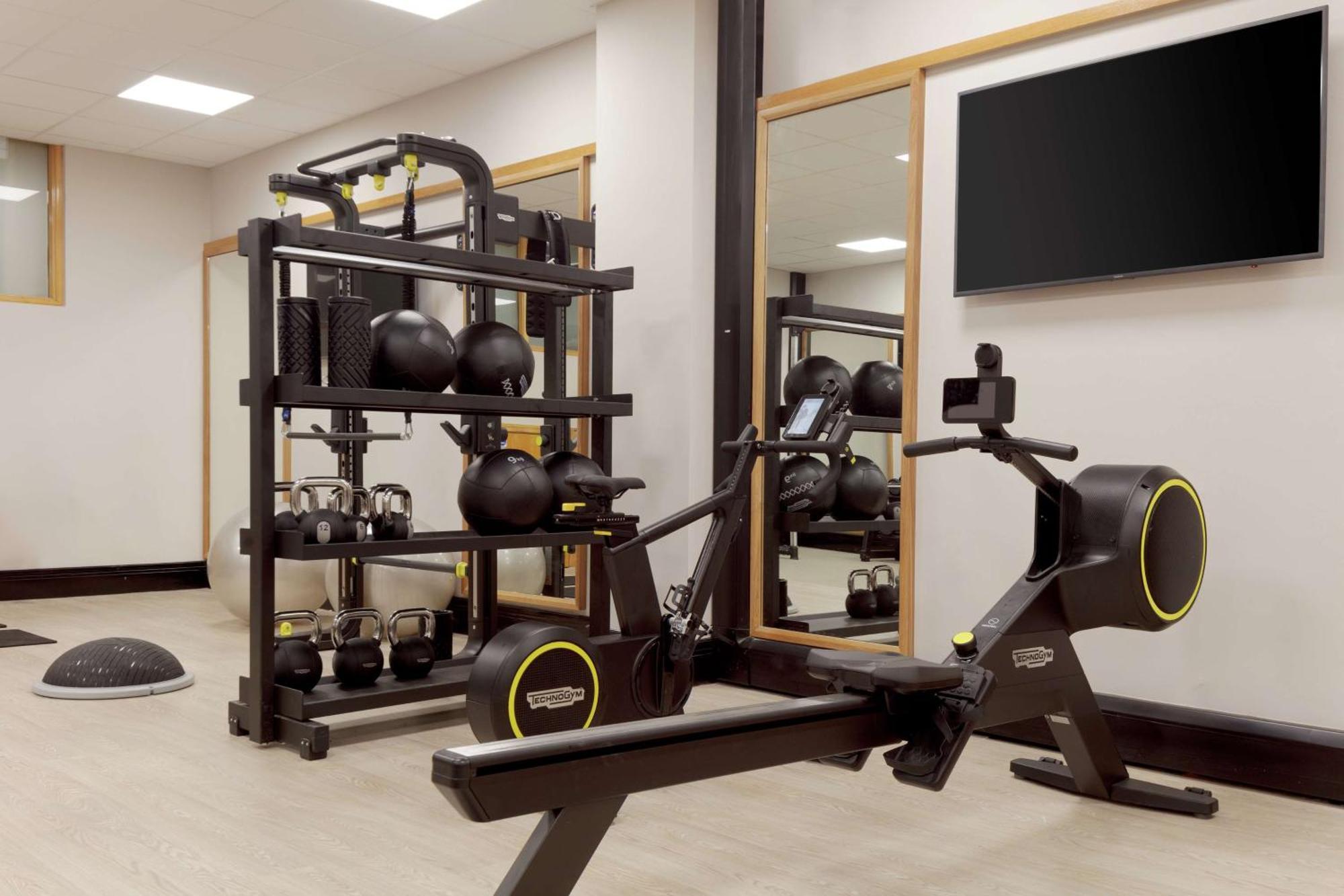Hilton Belfast Hotel Екстериор снимка The photo shows a fitness area featuring various exercise equipment. On the left side, there is a rack that holds multiple black kettlebells and a few other fitness accessories. In the center, there is a rowing machine designed for cardio workouts. T