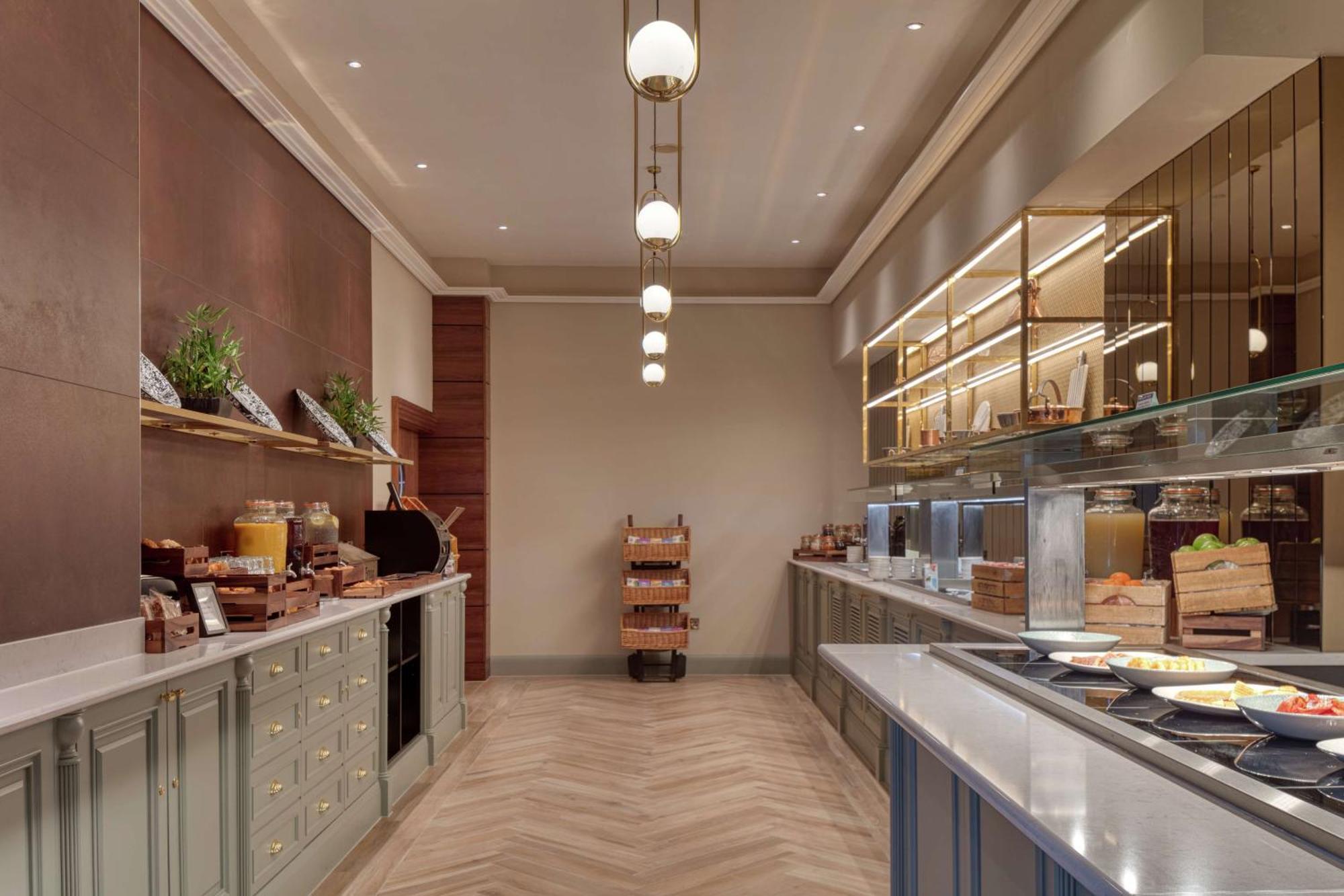 Hilton Belfast Hotel Екстериор снимка The photo shows an elegantly designed dining area, likely a buffet or breakfast setup. On one side, there are wooden shelves displaying various food items and plants, contributing to a warm atmosphere. Along the opposite side, there's a glass display