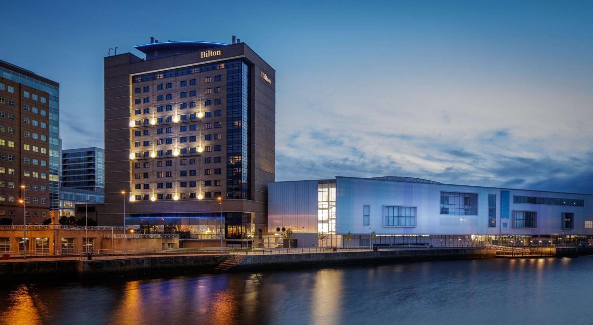 Hilton Belfast Hotel Екстериор снимка The image shows a Hilton hotel building along a waterway, illuminated during twilight. The hotel features a modern architectural design with multiple stories and large windows. To the right of the hotel, there is another structure that appears to be 
